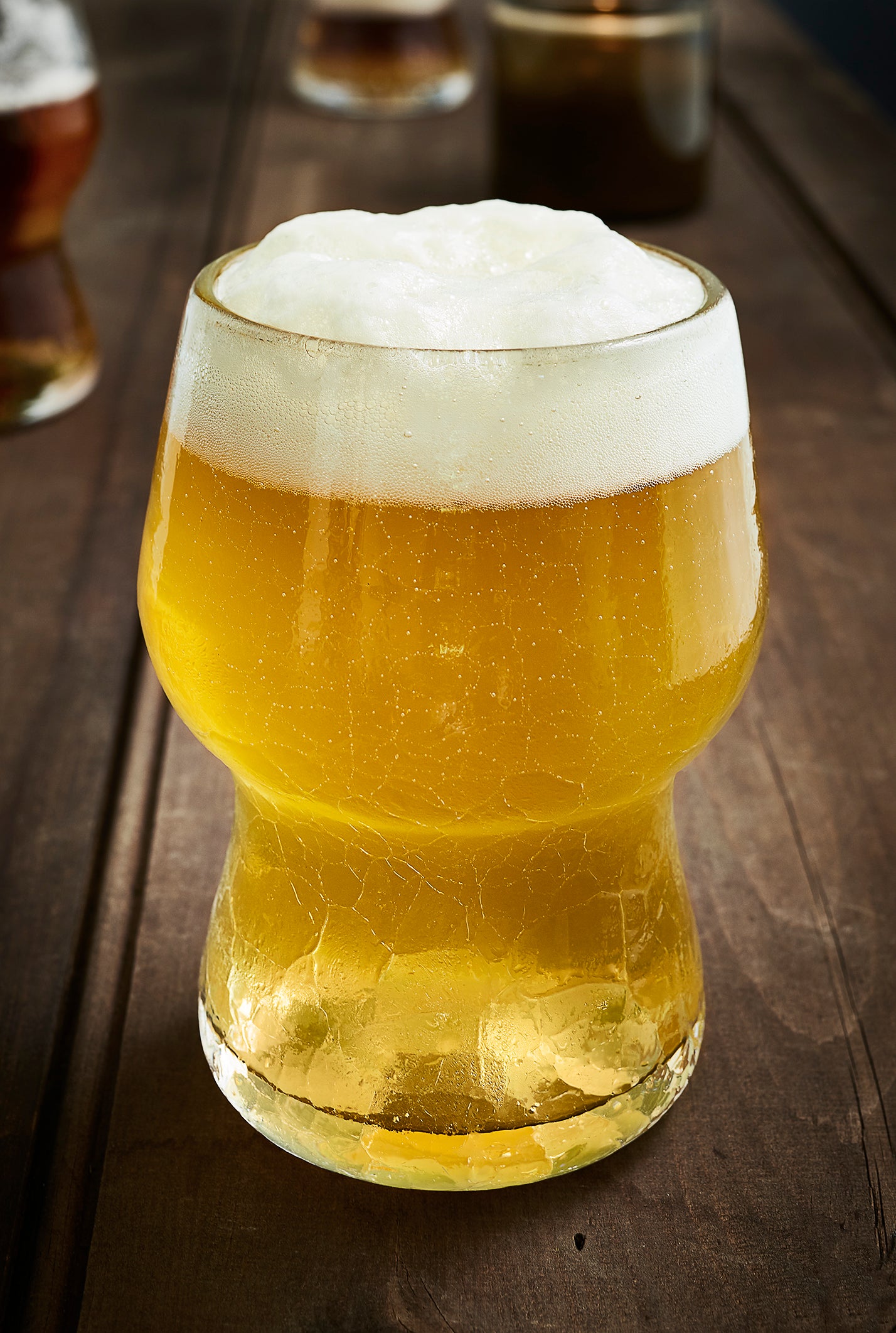 A clear Gray Art Glass crackle beer glass filled with beer sits on a wooden table.