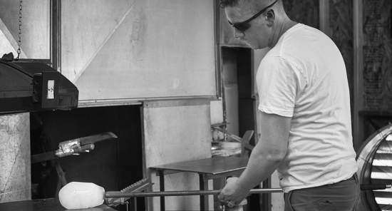 Glassblower and artist Michael Gray holding a pipe with a gather of molten glass on the end in his Gray Art Glass hot shop.