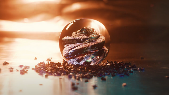 A round, clear glass paperweight with swirls of green and blue glass and cremation ash inside sit on a marver. Glass frit is scattered around the paperweight, illuminated by the soft glow of light from the glory hole.